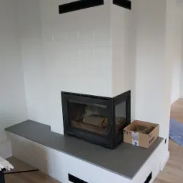Obudowa kominka wykończona białymi, strukturalnymi płytkami ceramicznymi z czarnymi akcentami, szarym blatem i wkładem kominkowym z palącym się drewnem wewnątrz. Obok stoi kartonowe pudełko.