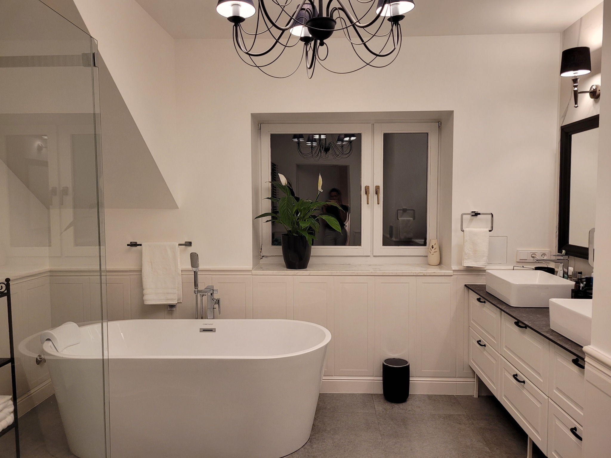 Elegancka łazienka w bieli i szarościach z wolnostojącą wanną, umywalkami nablatowymi i dekoracyjnym żyrandolem odbijającym się w oknie.