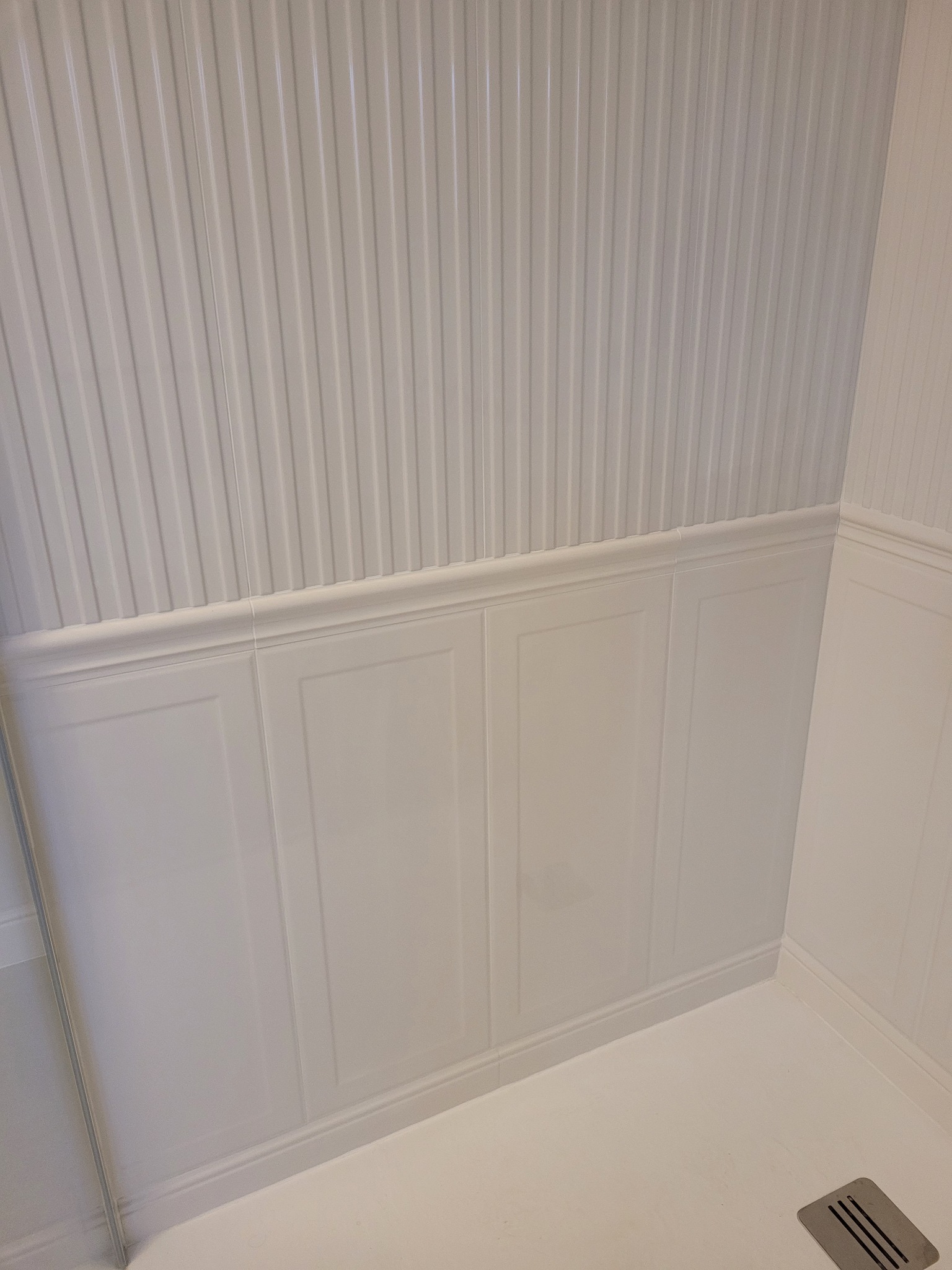 Biała łazienka z panelami ściennymi imitującymi boazerię angielską, wykończona listwami przypodłogowymi i dekoracyjnymi, z odpływem liniowym w podłodze.