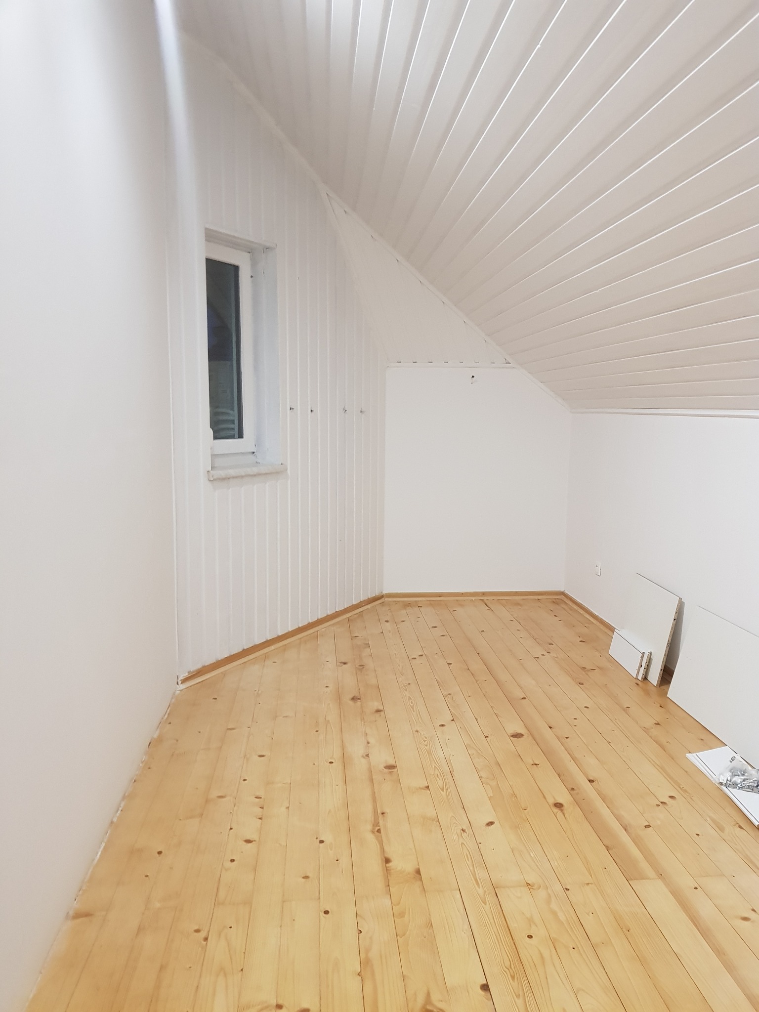 Poddasze z drewnianą podłogą i białymi ścianami wykończonymi panelami, widoczne okno i białe elementy meblowe w rogu.
