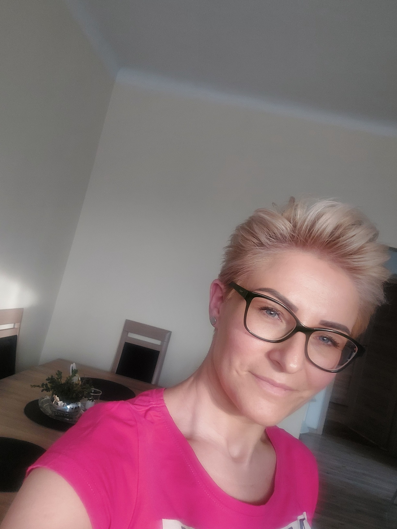 Uśmiechnięta kobieta w okularach i różowej koszulce, w tle stół z dekoracją roślinną i krzesła, jasne wnętrze.