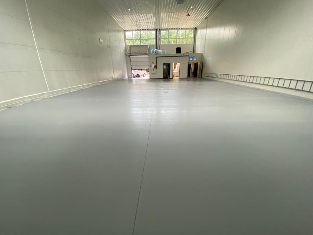 Szara, gładka podłoga w hali magazynowej, widoczna brama wjazdowa i drabina oparta o ścianę.