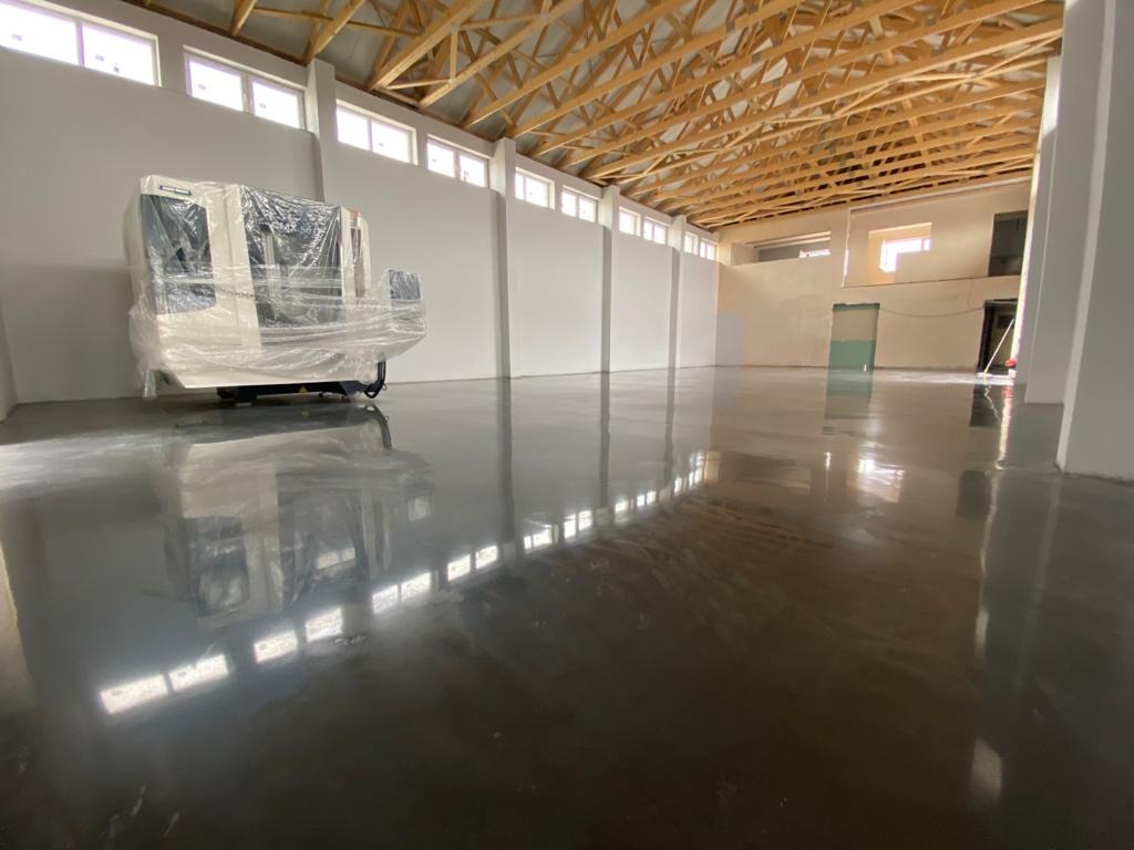 Błyszcząca, ciemnoszara podłoga w hali przemysłowej, z maszyną w folii ochronnej w tle i drewnianą konstrukcją dachu.