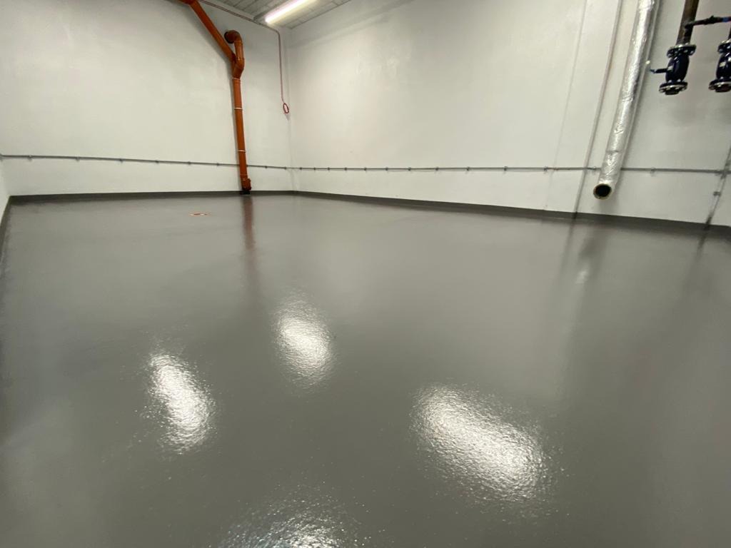 Gładka, szara podłoga w przemysłowym pomieszczeniu z rurami i instalacjami na ścianach.