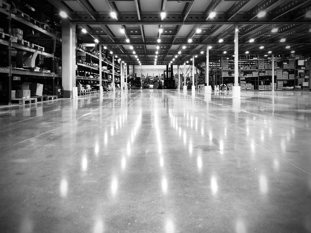Wnętrze magazynu z polerowaną, bezszwową posadzką, odbijającą światła lamp, w tle widoczne regały i wózki widłowe, zdjęcie czarno-białe.