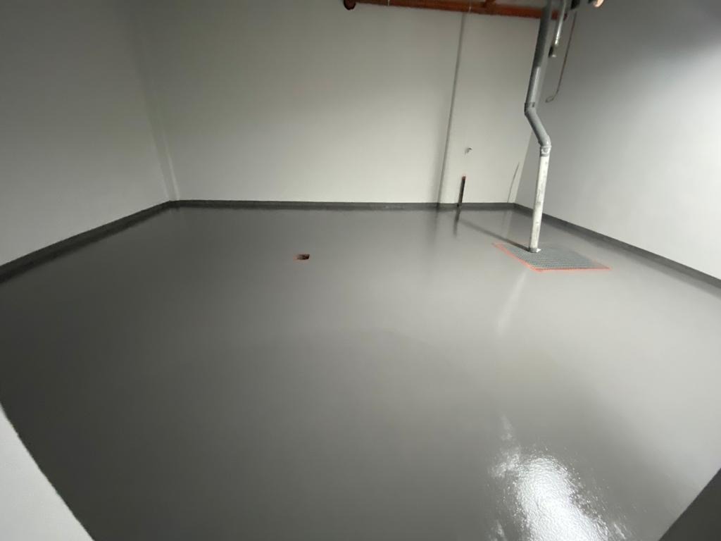 Szara, gładka posadzka w pomieszczeniu przemysłowym, z widocznym odpływem liniowym i rurą spustową. Minimalistyczne wykończenie, jednolite, jasne ściany.