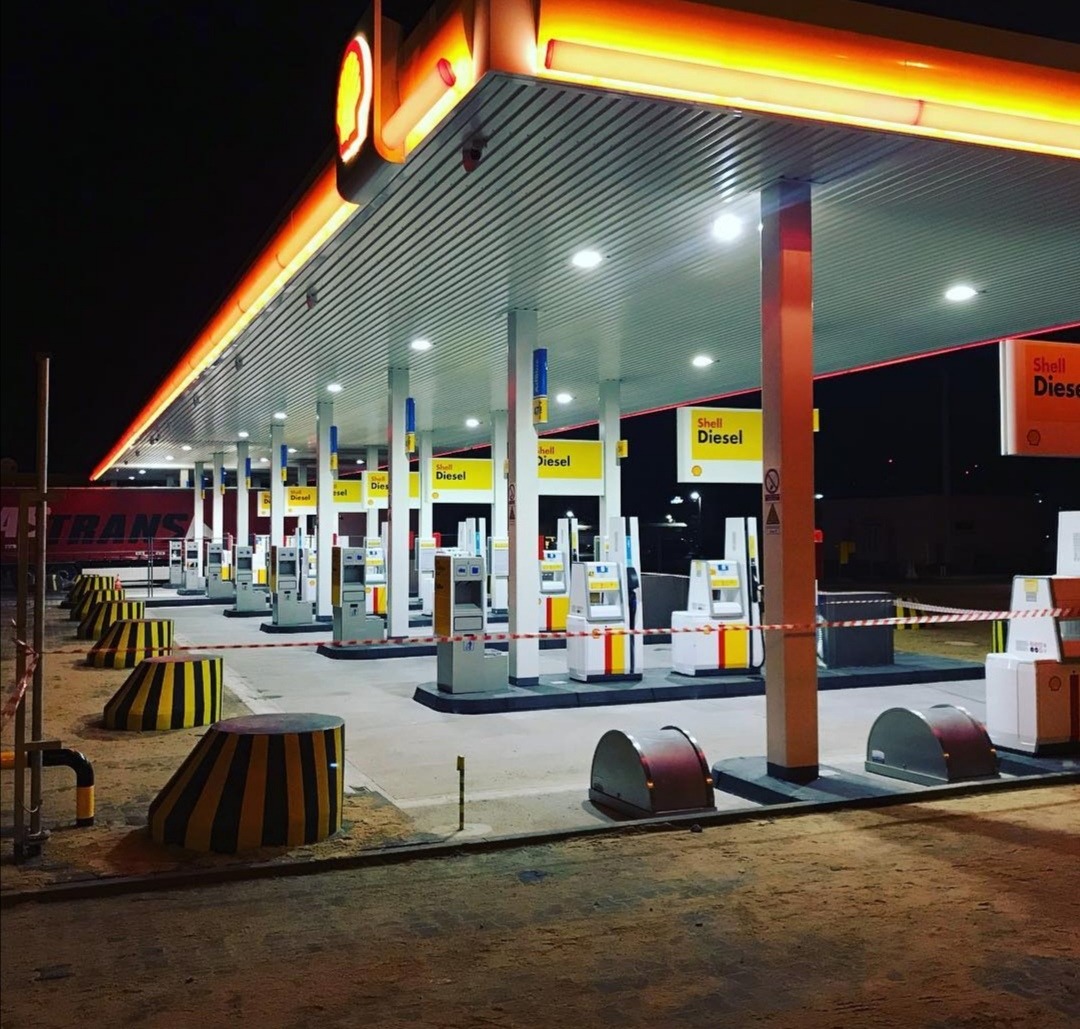 Oświetlona stacja benzynowa Shell nocą, z dystrybutorami paliwa i betonowymi barierami w czarno-żółte pasy.