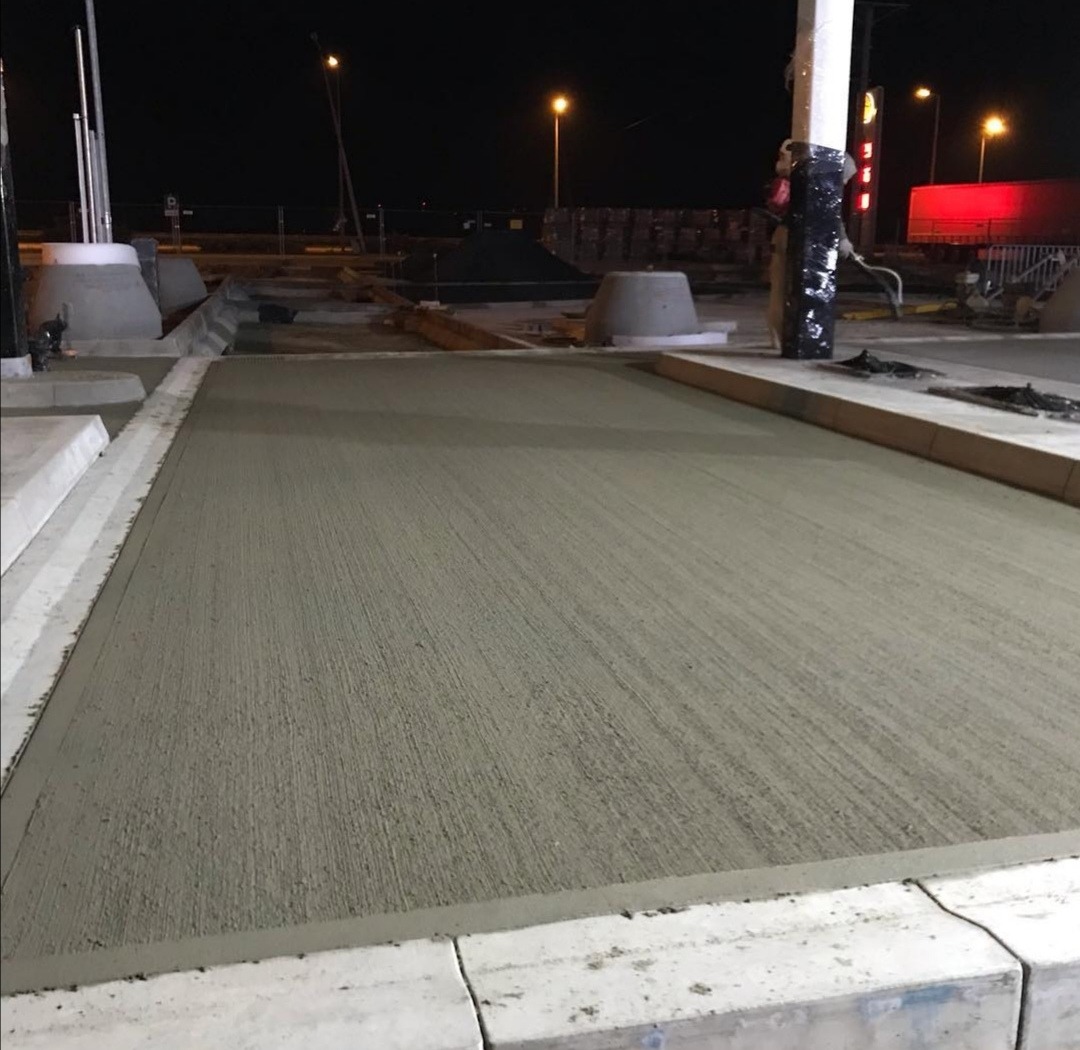 Świeżo wylana, szara powierzchnia jastrychu na zewnątrz, otoczona betonowymi krawężnikami, widoczna nocą w świetle latarni, w tle elementy budowy.