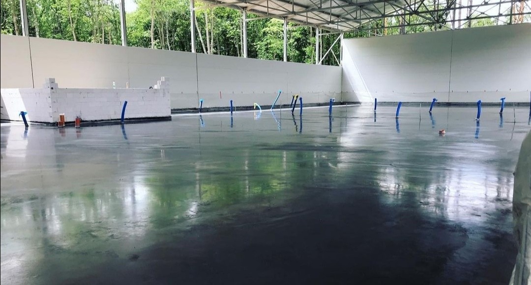 Wylewka samopoziomująca w hali przemysłowej, widoczne wystające rurki instalacyjne i fragment murku z bloczków betonowych przy ścianie