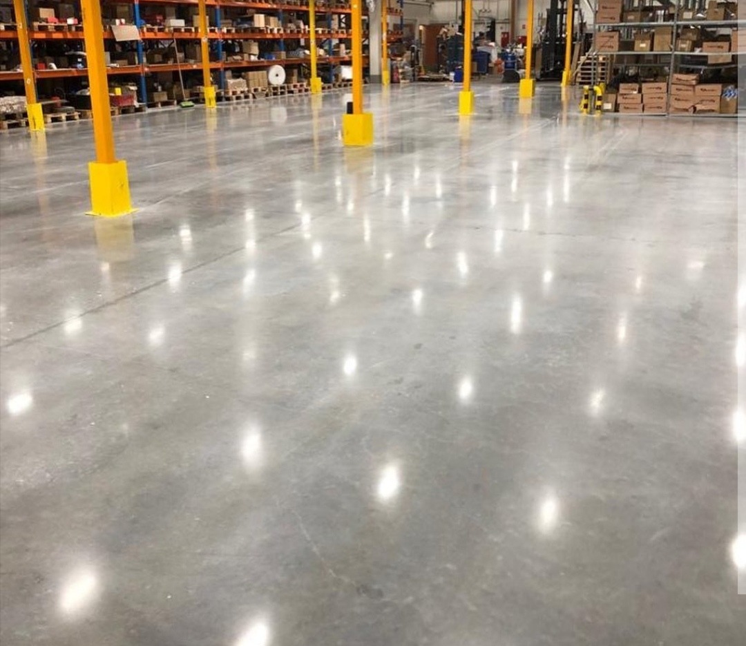 Błyszcząca, gładka posadzka przemysłowa w hali magazynowej w Bydgoszczy, z żółtymi słupami zabezpieczającymi regały, odbijająca światło lamp sufitowych.