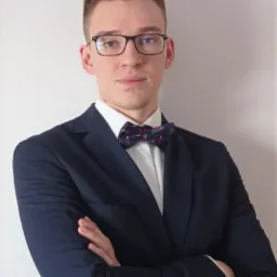 Portret młodego mężczyzny w okularach, granatowej marynarce i ozdobnej muszce, z założonymi rękami na tle jasnej ściany.