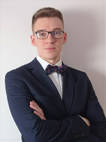 Portret młodego mężczyzny w okularach, granatowej marynarce i ozdobnej muszce, z założonymi rękami na tle jasnej ściany.