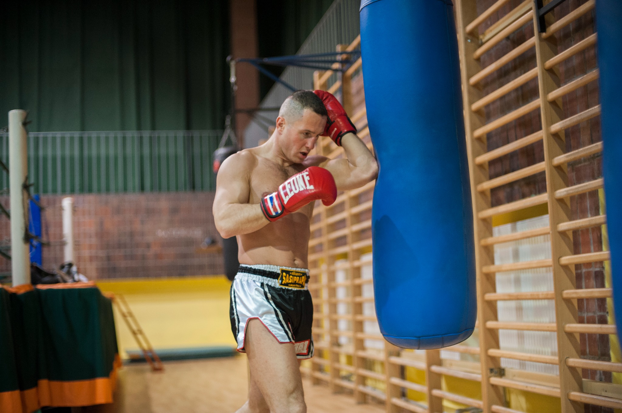Mężczyzna bez koszulki w czerwonych rękawicach bokserskich, w spodenkach do muay thai, opiera się o niebieski worek treningowy na tle drabinek gimnastycznych w sali treningowej.