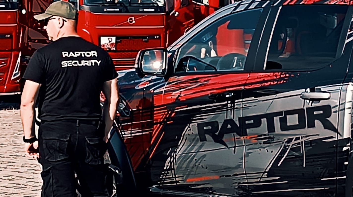 Mężczyzna w czarnej koszulce z napisem 'RAPTOR SECURITY' stoi obok pickupa z graficznym wzorem i napisem 'RAPTOR', w tle czerwone ciężarówki Volvo.