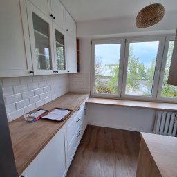 Remonty i Wykończenia Łukasz Grochecki - Jasna kuchnia z białymi szafkami, drewnianym blatem i podłogą, okno z widokiem na zieleń, lampa z wikliny na suficie, płytki imitujące cegłę na ścianie.