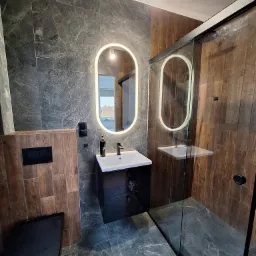 Nowoczesna łazienka z czarnymi akcentami: umywalka z szafką, podświetlane lustro, kabina prysznicowa i wisząca toaleta, wykończona płytkami imitującymi marmur i drewno.