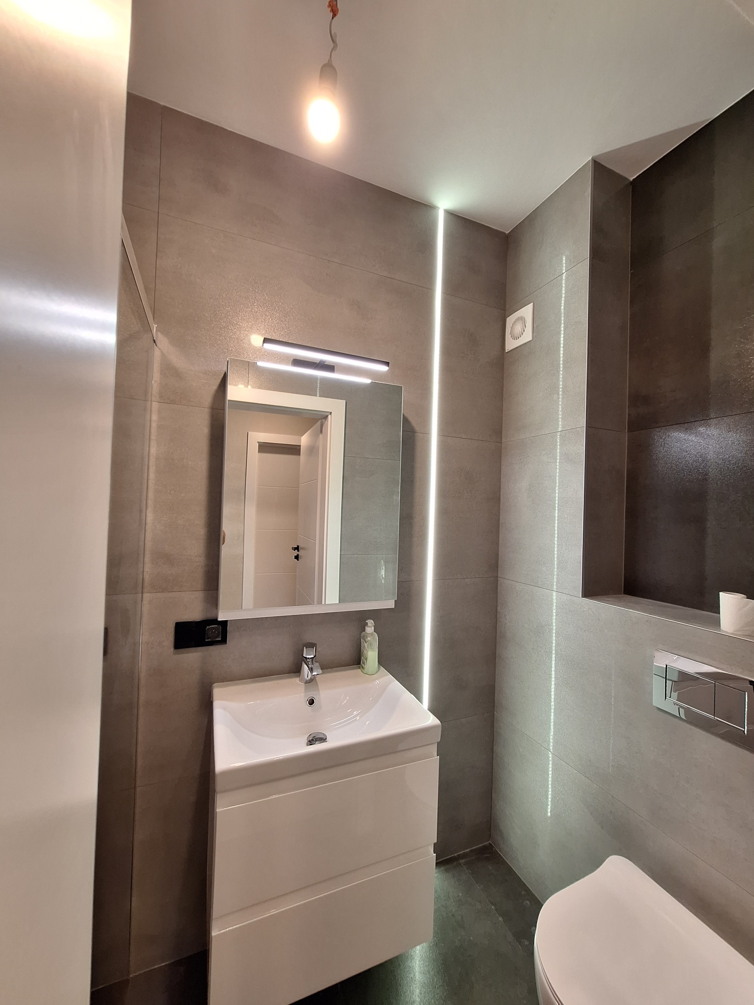 Nowoczesna łazienka z szarymi płytkami, białym umywalką z szafką, podświetlanym lustrem i pionowym oświetleniem LED.