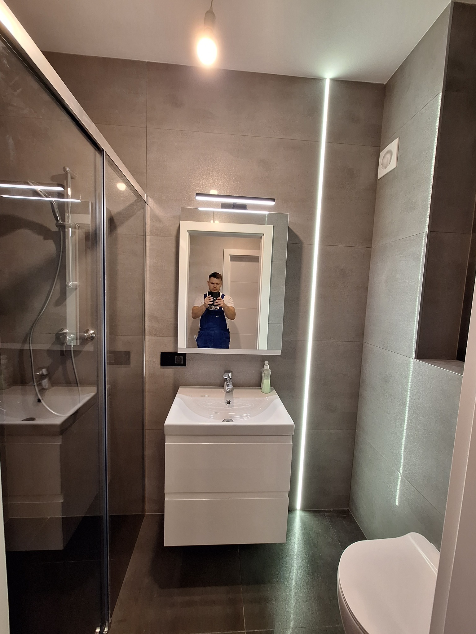 Wykończona łazienka w szarościach z kabiną prysznicową, umywalką z szafką i toaletą, oświetlona listwą LED w pionowym wyżłobieniu w ścianie. W lustrze odbicie fotografa w niebieskim kombinezonie.