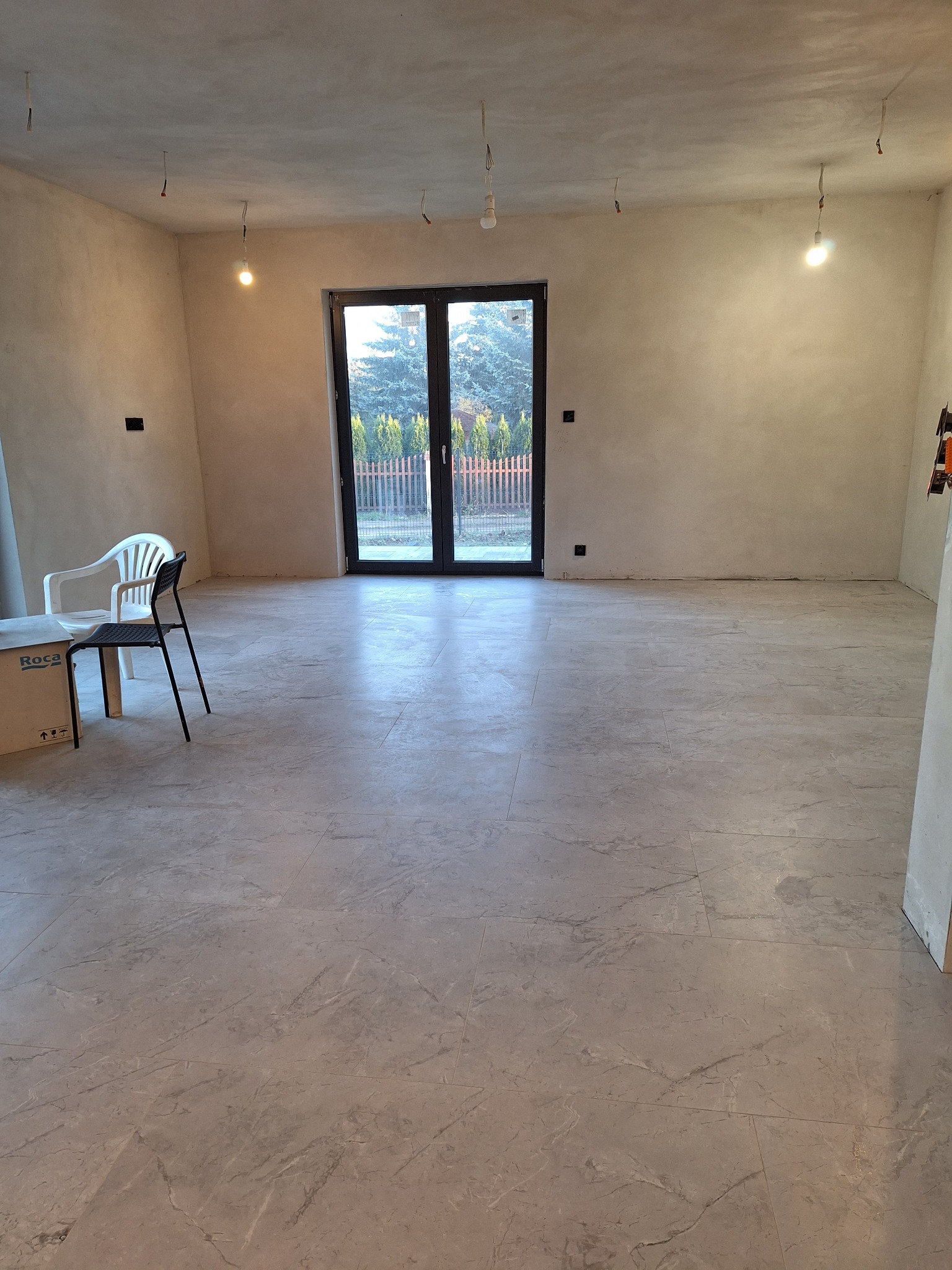 Wykończone wnętrze: Jasnoszara podłoga z płytek imitujących marmur, ściany po szpachlowaniu, widoczne zawieszone kable od oświetlenia i okno balkonowe na tle zieleni.