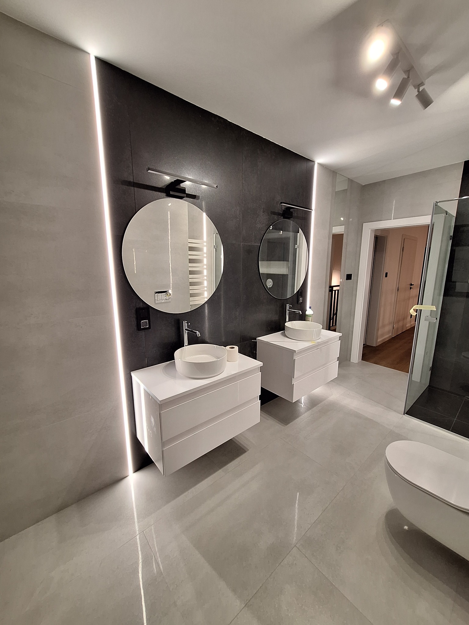 Nowoczesna łazienka z dwoma umywalkami nablatowymi na białych szafkach, okrągłe lustra z podświetleniem, szara płytka na ścianach i podłodze, oświetlenie punktowe na suficie.