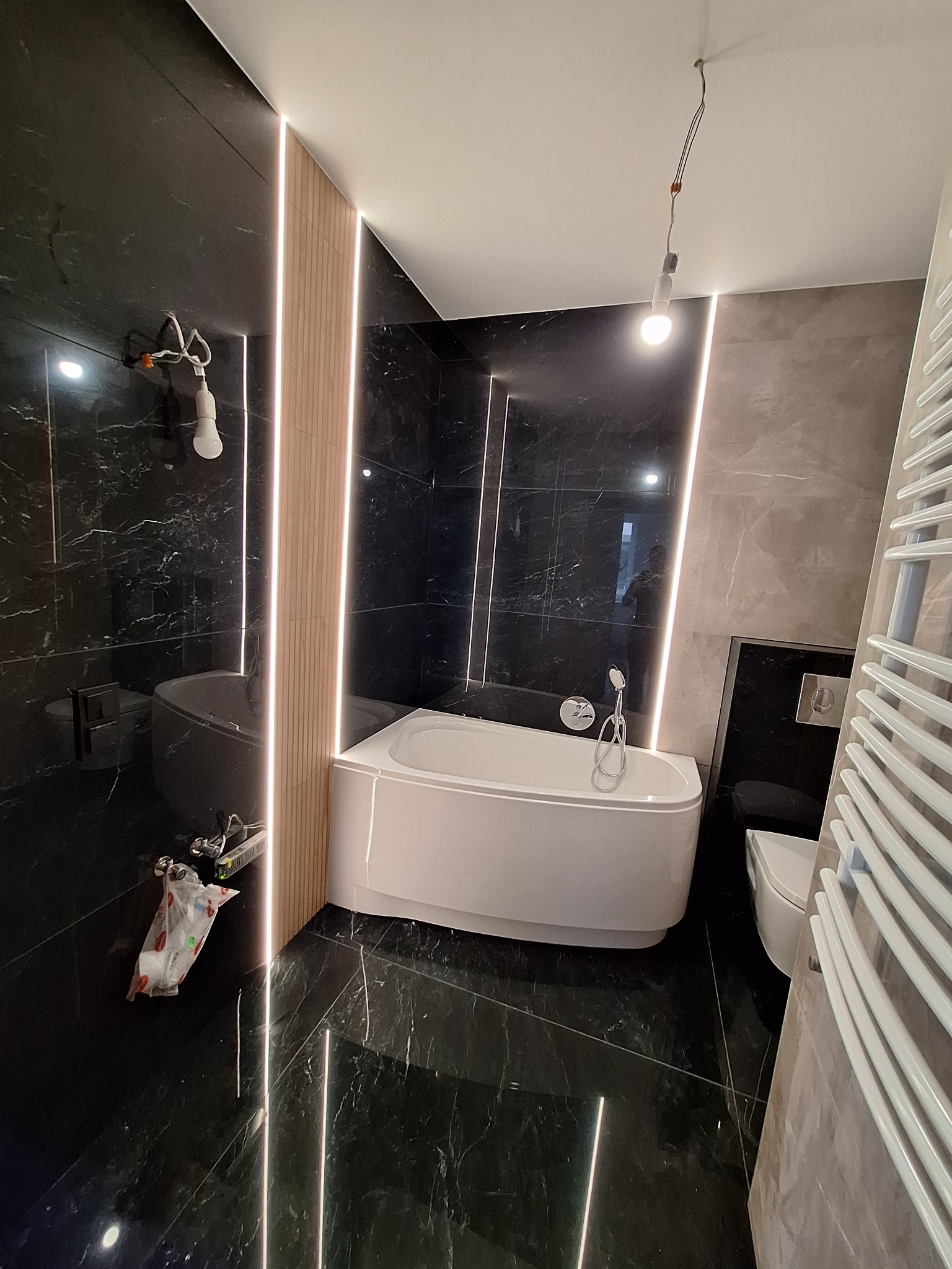 Wnętrze łazienki w trakcie wykańczania: czarne marmurowe płytki, biała wanna narożna, grzejnik drabinkowy, podwieszana toaleta, oświetlenie LED w pionowych pasach, wisząca żarówka na kablu.