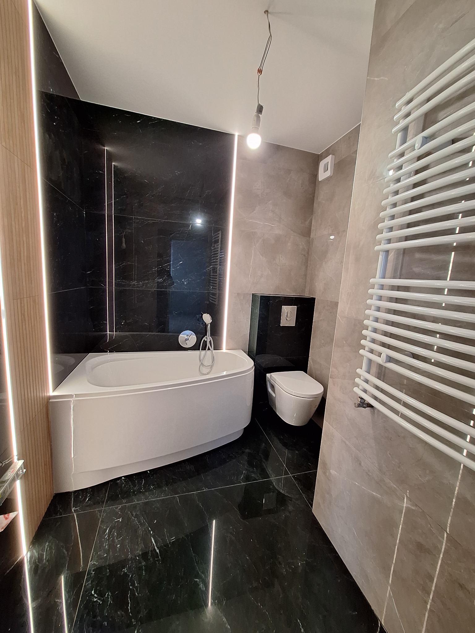 Nowoczesna łazienka z czarnymi marmurowymi płytkami, podświetlaną wanną narożną, wiszącą toaletą i grzejnikiem drabinkowym.