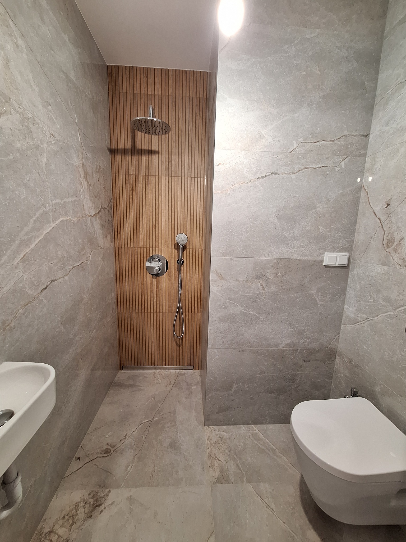 Nowoczesna łazienka z szarymi płytkami imitującymi marmur, drewnianą ścianą prysznicową z deszczownicą i minimalistyczną armaturą oraz podwieszaną toaletą.