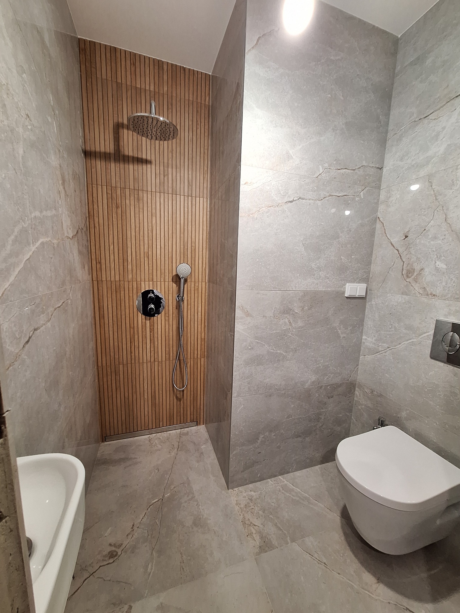 Nowoczesna łazienka z kabiną prysznicową wyłożoną drewnopodobnymi panelami, deszczownicą i baterią podtynkową, szare płytki imitujące marmur na ścianach i podłodze, wisząca toaleta i umywalka.