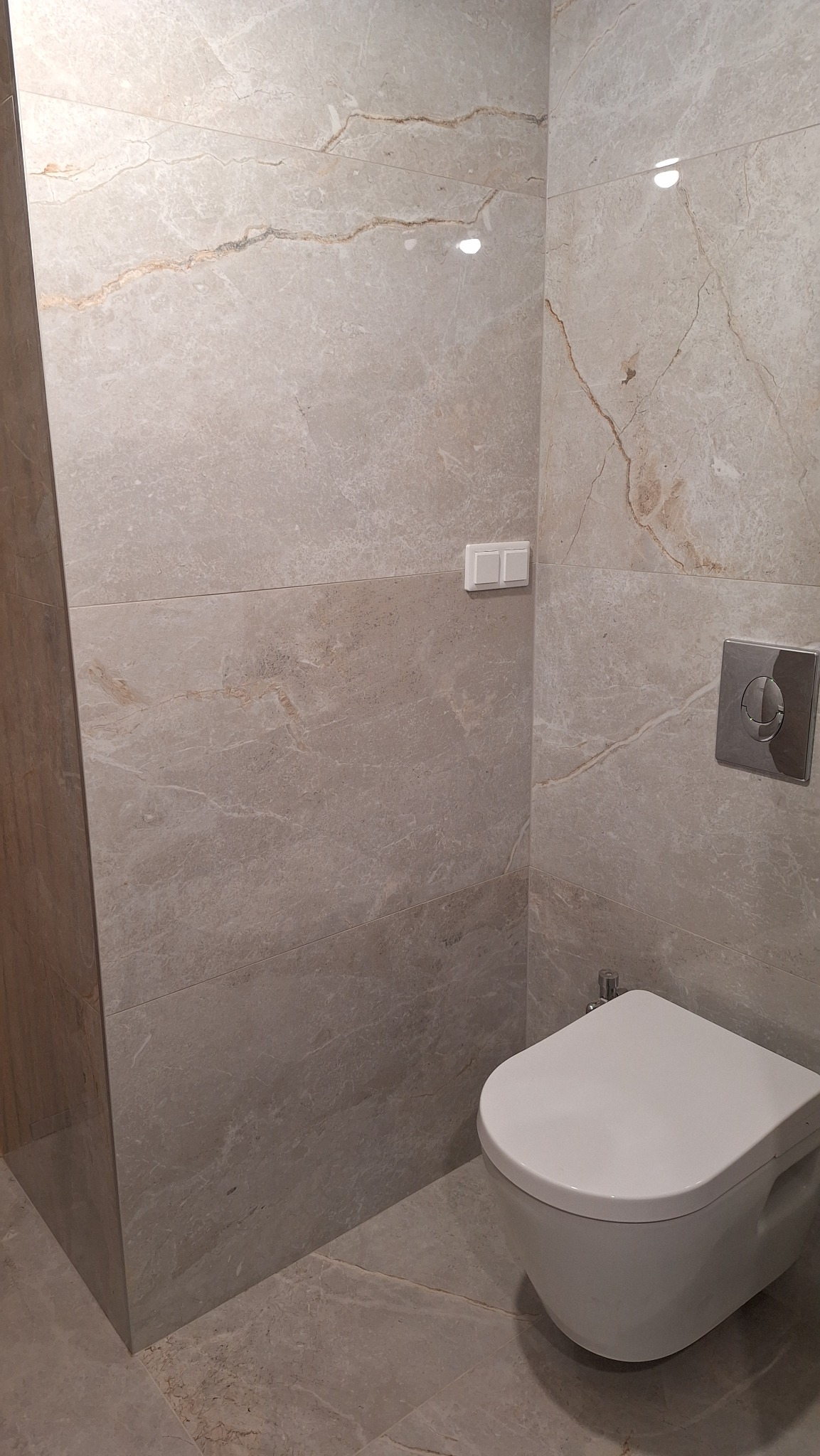 Nowoczesna toaleta podwieszana w łazience wykończonej płytkami imitującymi marmur w odcieniach beżu i szarości, z widocznym przyciskiem spłukującym i włącznikiem światła.