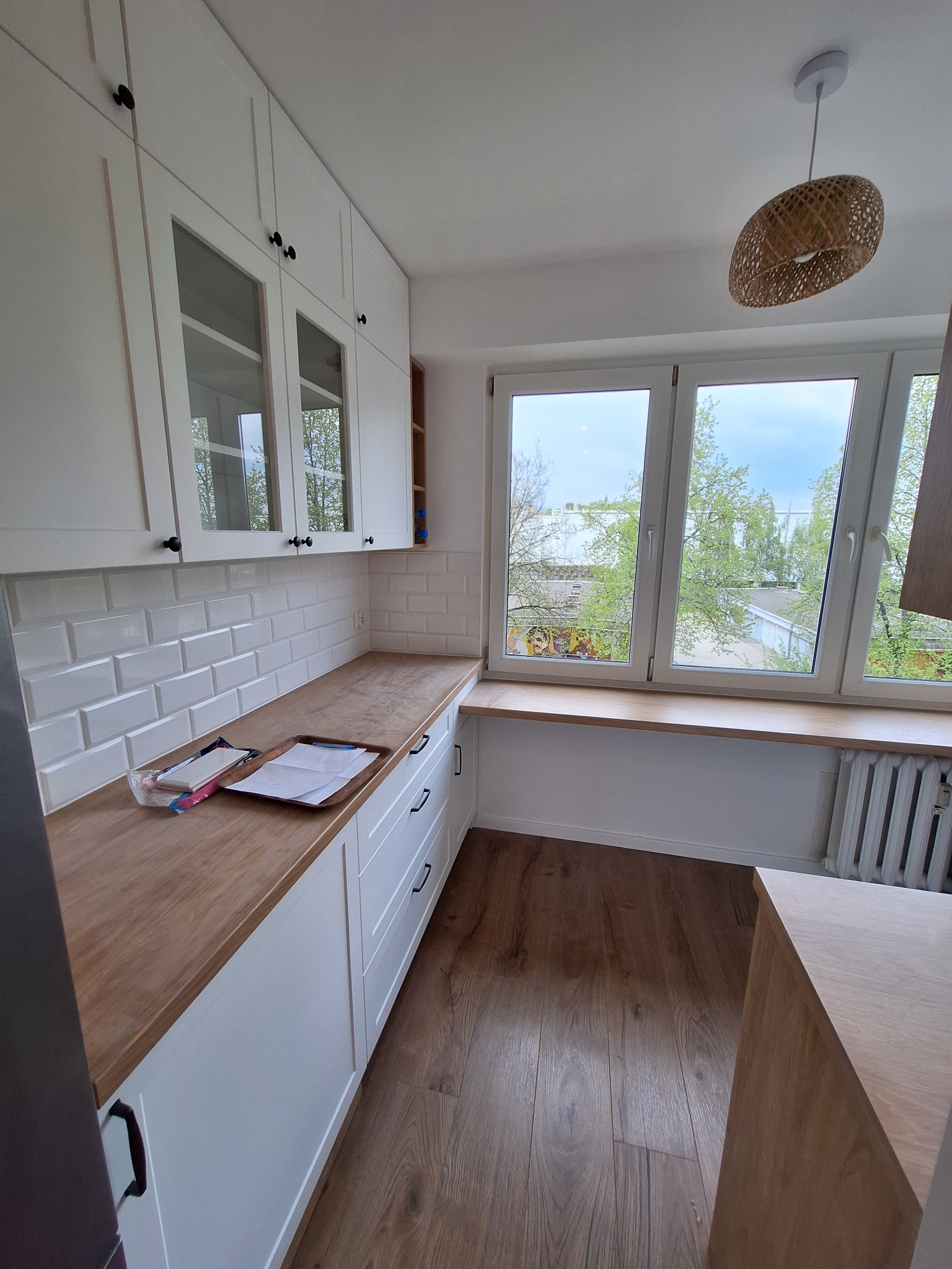 Jasna kuchnia z białymi szafkami, drewnianym blatem i podłogą, okno z widokiem na zieleń, lampa z wikliny na suficie, płytki imitujące cegłę na ścianie.