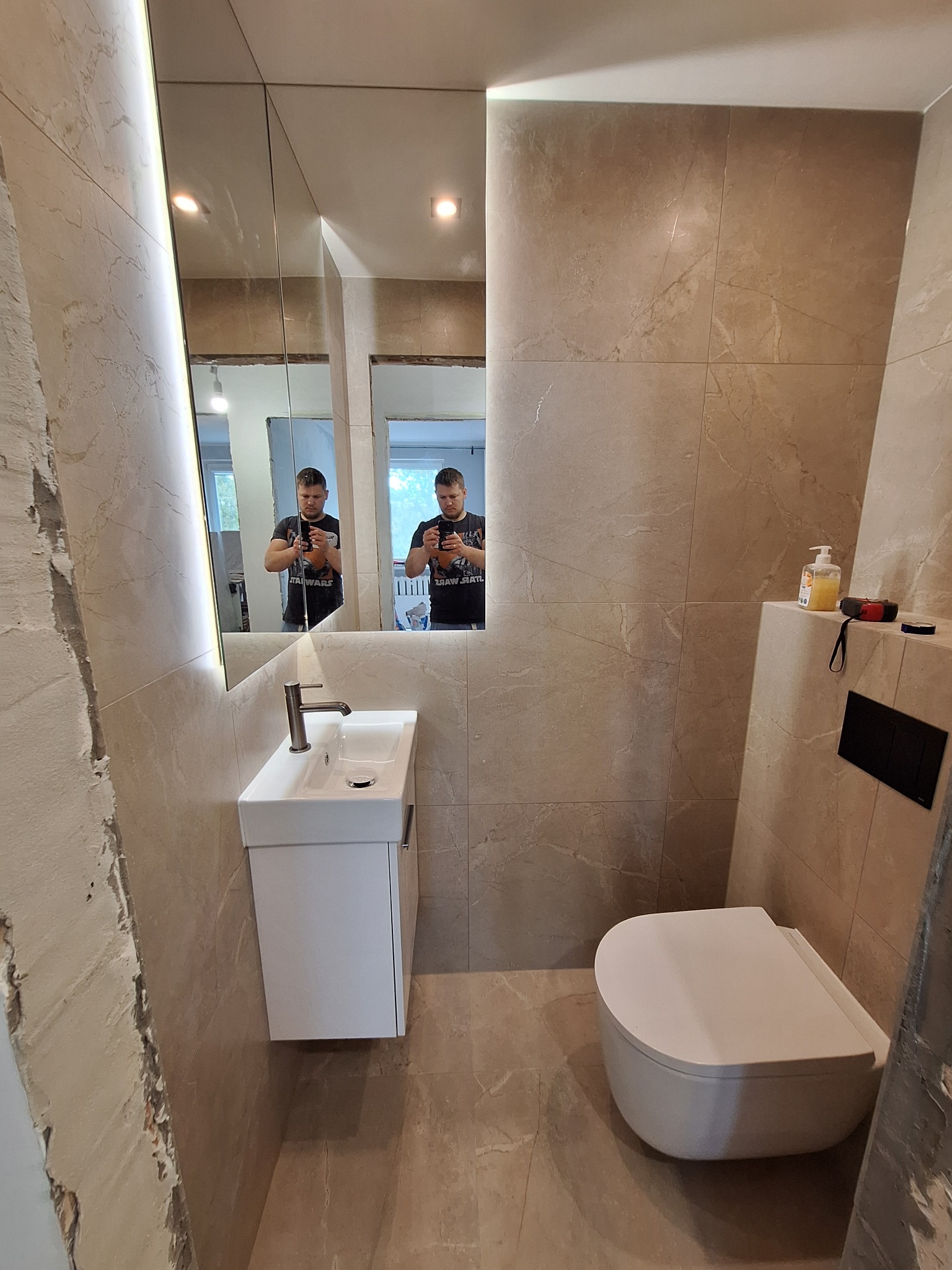 Wykończona łazienka z beżowymi płytkami, podwieszaną toaletą i umywalką z szafką; w lustrze odbija się osoba robiąca zdjęcie.