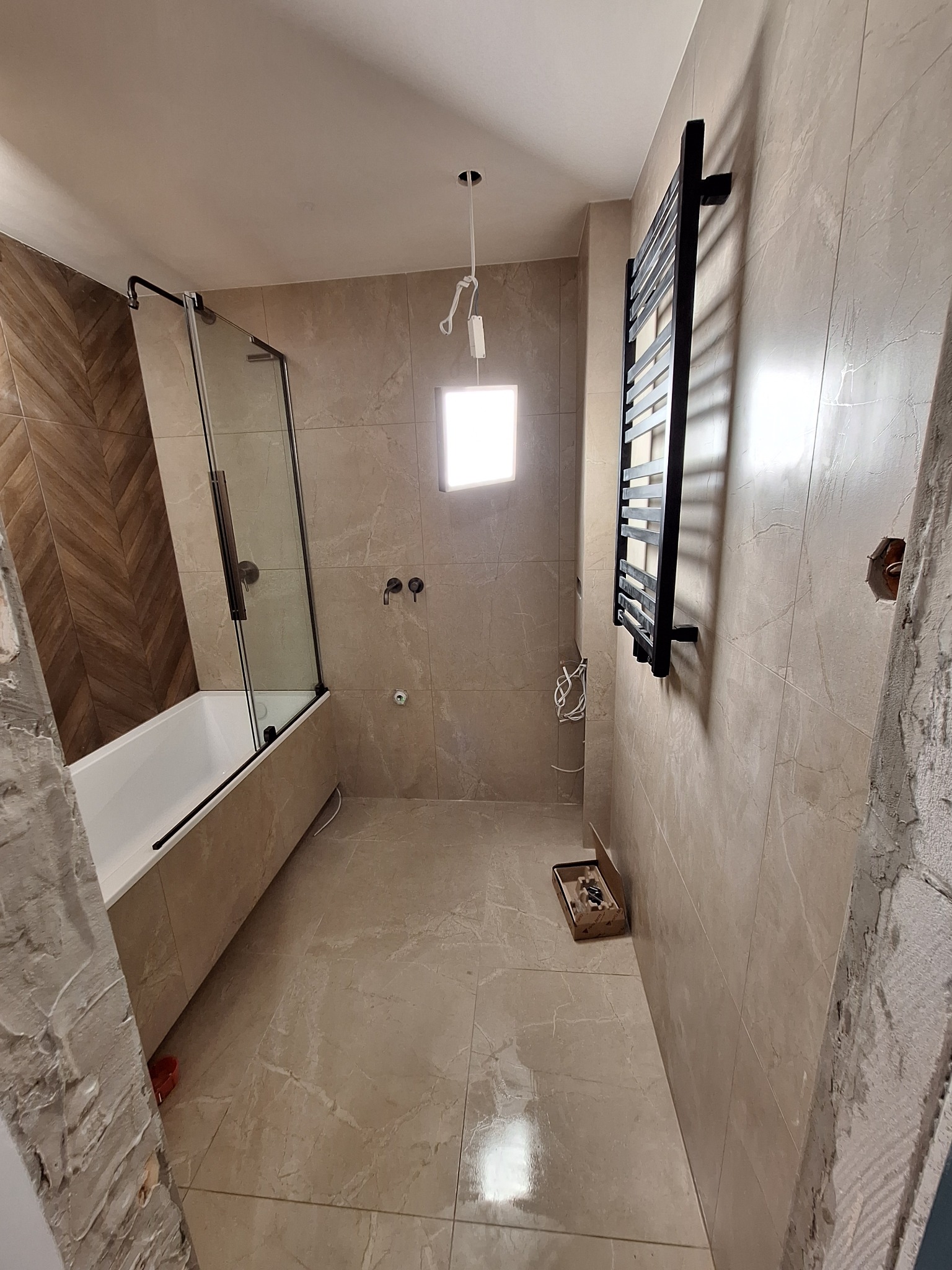 Łazienka w trakcie remontu: zamontowana kabina prysznicowa z czarnymi elementami, grzejnik drabinkowy na ścianie z beżowych płytek, widoczne niedokończone obrzeża i wystające kable.