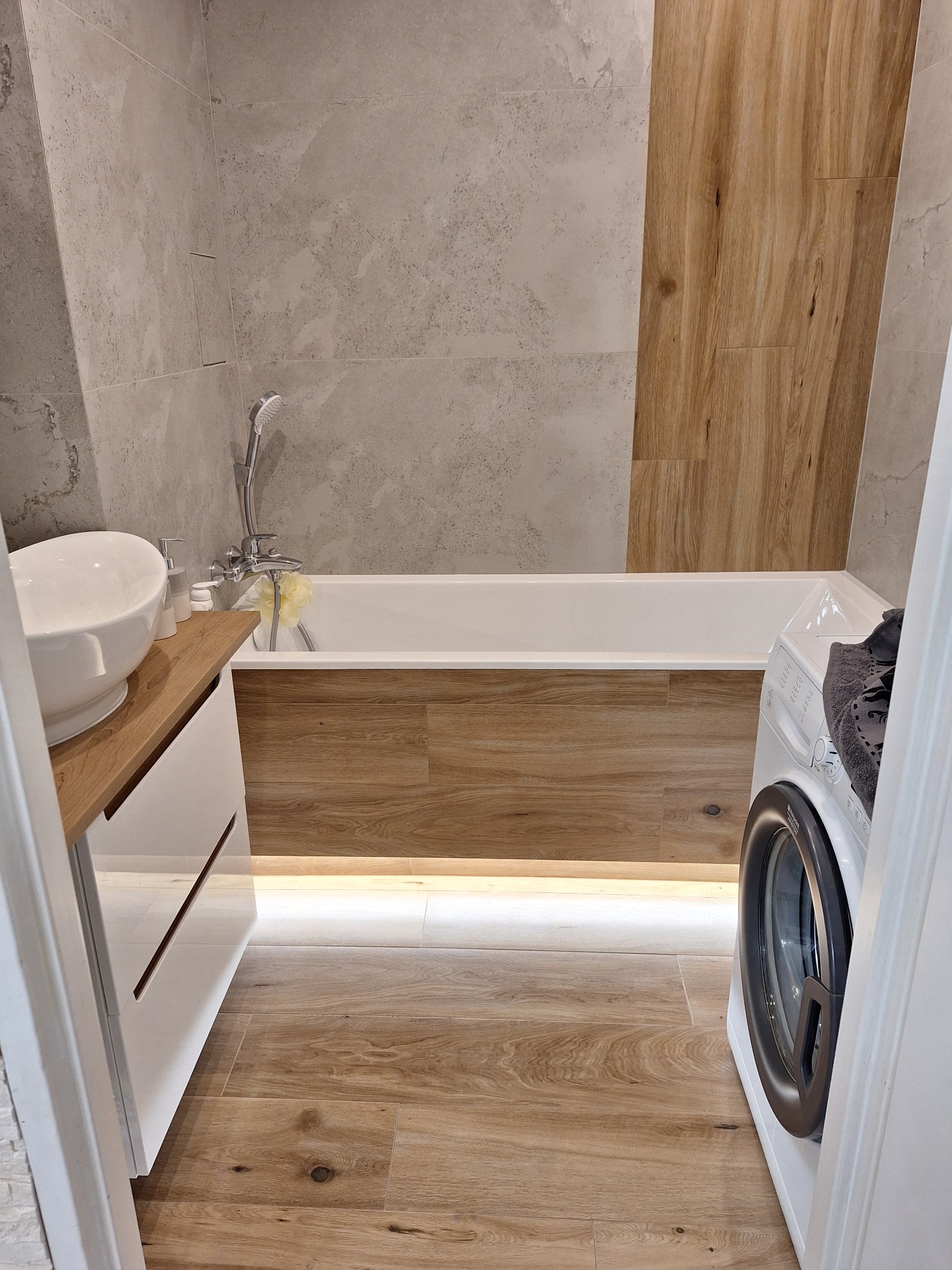 Nowoczesna łazienka z drewnopodobnymi płytkami na podłodze i obudowie wanny, umywalką nablatową, pralką i podświetleniem LED pod wanną.