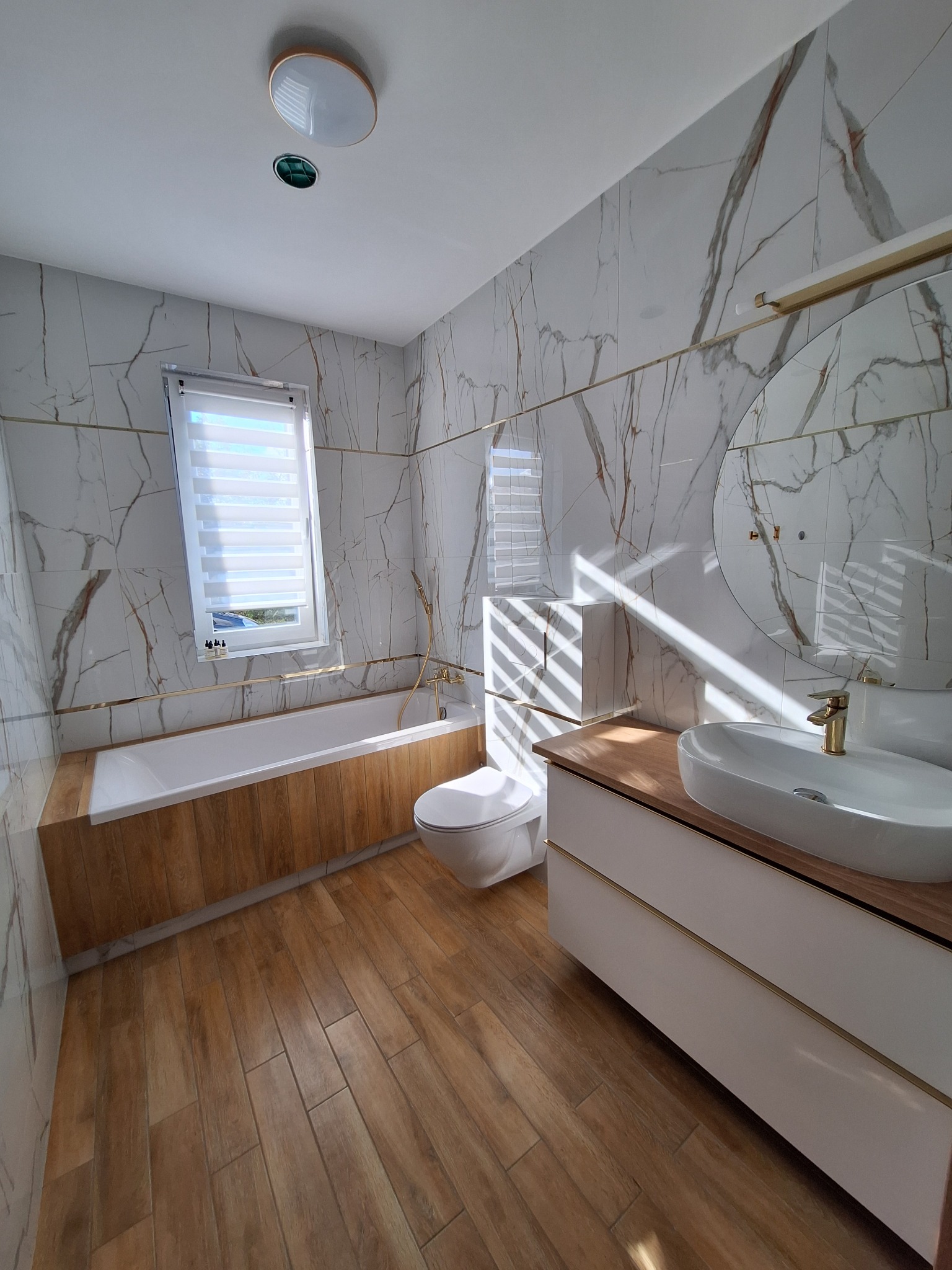 Nowoczesna łazienka z marmurowymi płytkami, drewnopodobną zabudową wanny i podłogą, owalnym lustrem i białą szafką z drewnianym blatem.