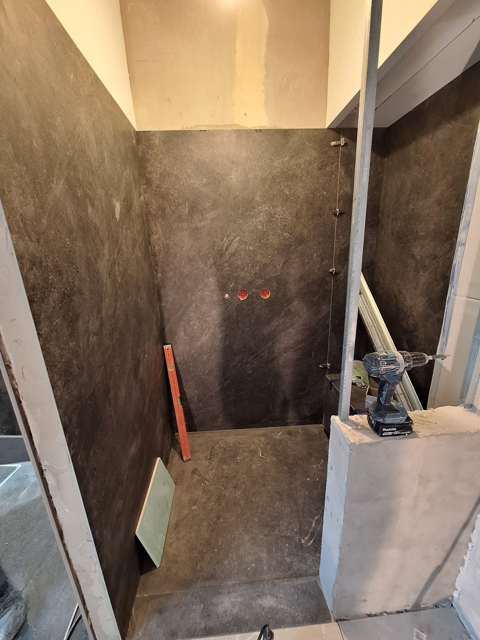 Montaż kabiny prysznicowej z ciemnymi płytkami, widoczny poziomica, wiertarka Makita i profile metalowe do mocowania szkła.