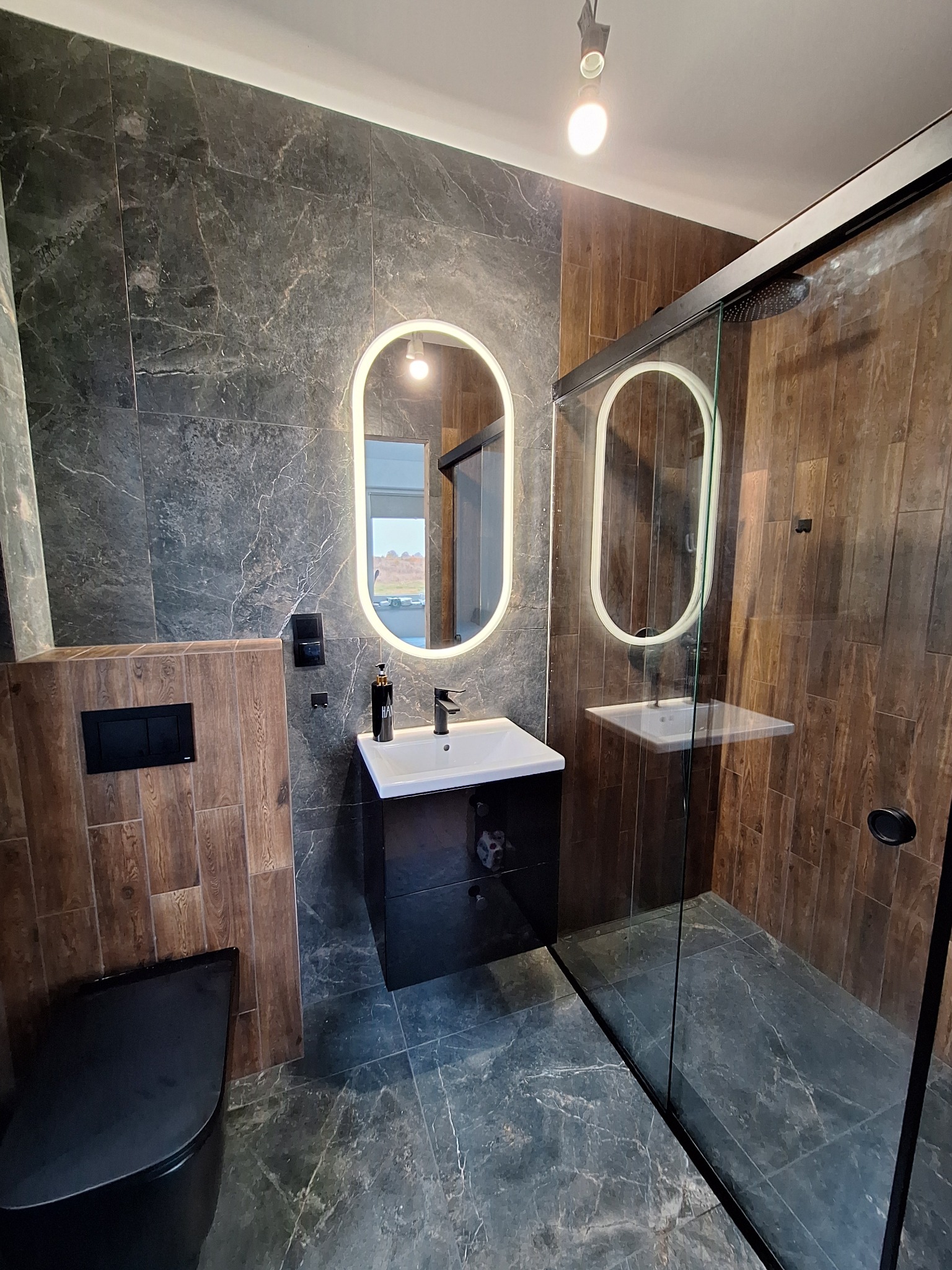 Nowoczesna łazienka z czarnymi akcentami: umywalka z szafką, podświetlane lustro, kabina prysznicowa i wisząca toaleta, wykończona płytkami imitującymi marmur i drewno.