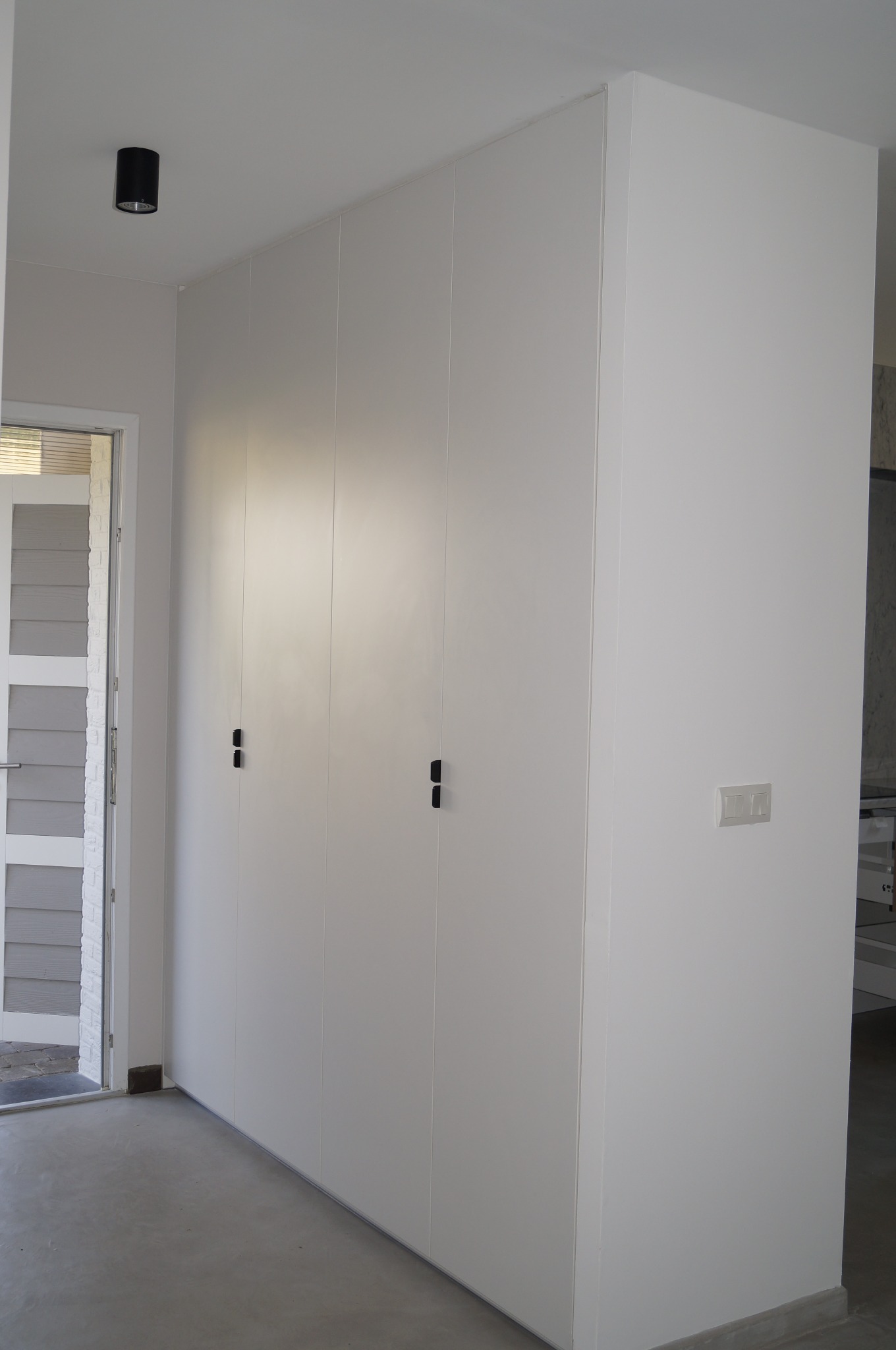 Wysoka, biała szafa wnękowa z czterema drzwiami i minimalistycznymi, czarnymi uchwytami, w jasnym pomieszczeniu z betonową podłogą.