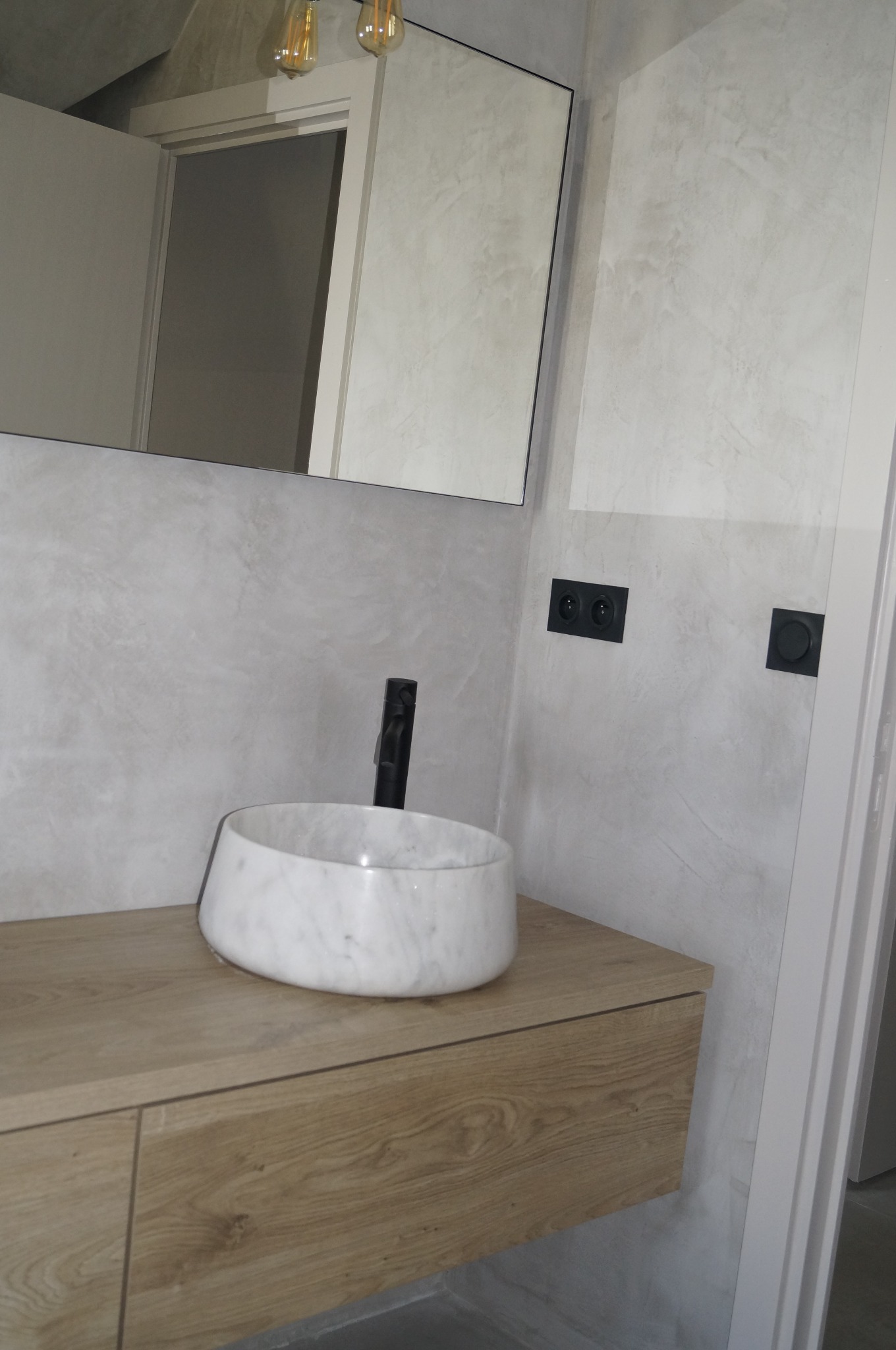 Nowoczesna łazienka z marmurową umywalką nablatową na drewnianej szafce, ściany wykończone w odcieniach szarości z widocznymi gniazdkami elektrycznymi, minimalistyczna armatura w kolorze czarnym...
