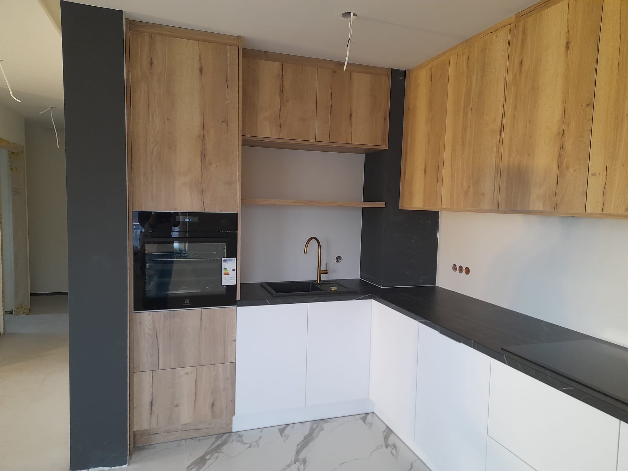 Nowoczesna kuchnia z drewnianymi frontami szafek górnych i dolnych, białymi frontami szafek dolnych, czarnym blatem i złotą baterią, widok na marmurową podłogę i częściowo wykończone ściany.