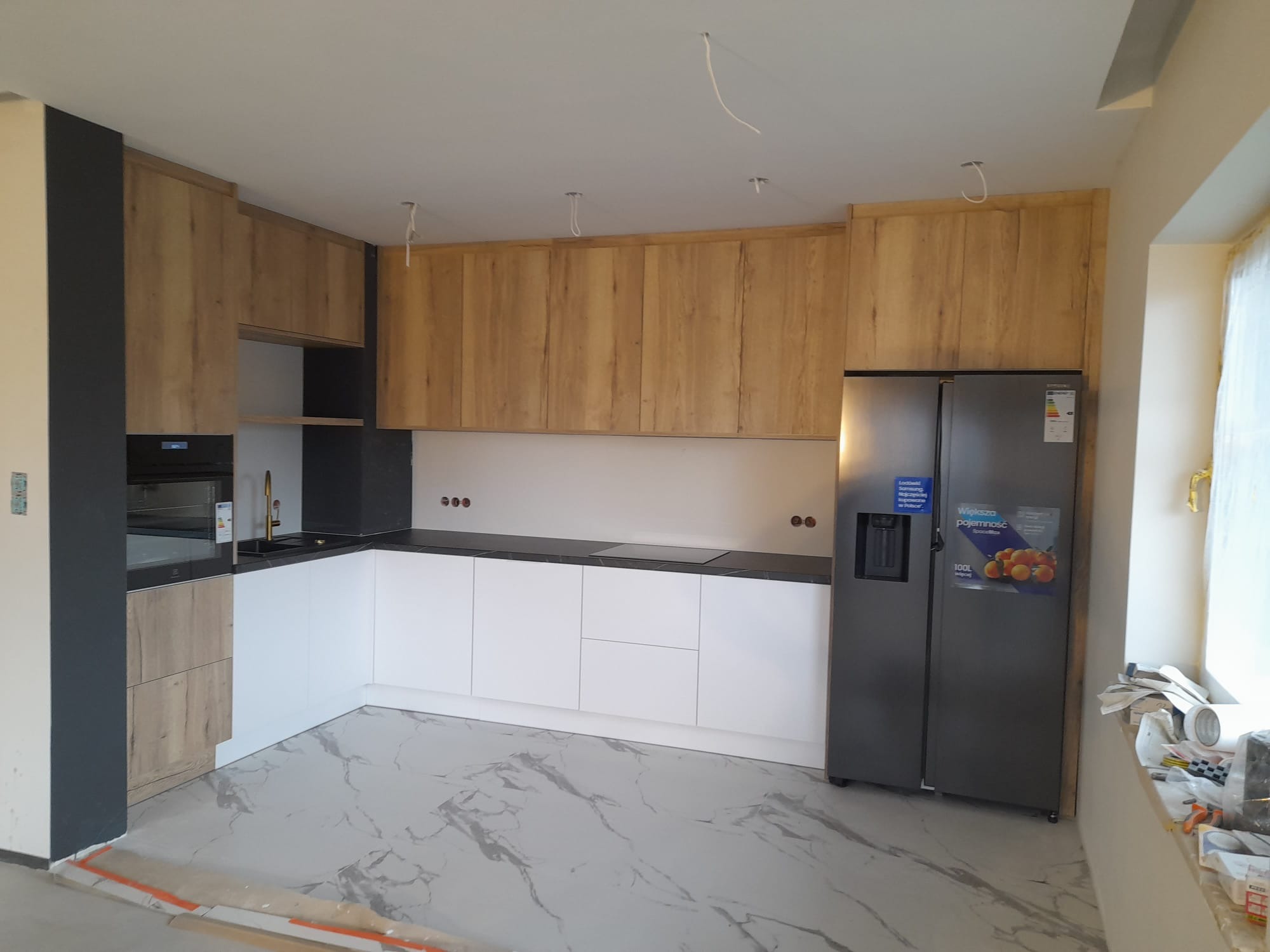 Nowoczesna kuchnia z frontami w kolorze jasnego drewna i białymi szafkami dolnymi, czarny blat, marmurowe płytki podłogowe, widoczne wystające kable od oświetlenia.
