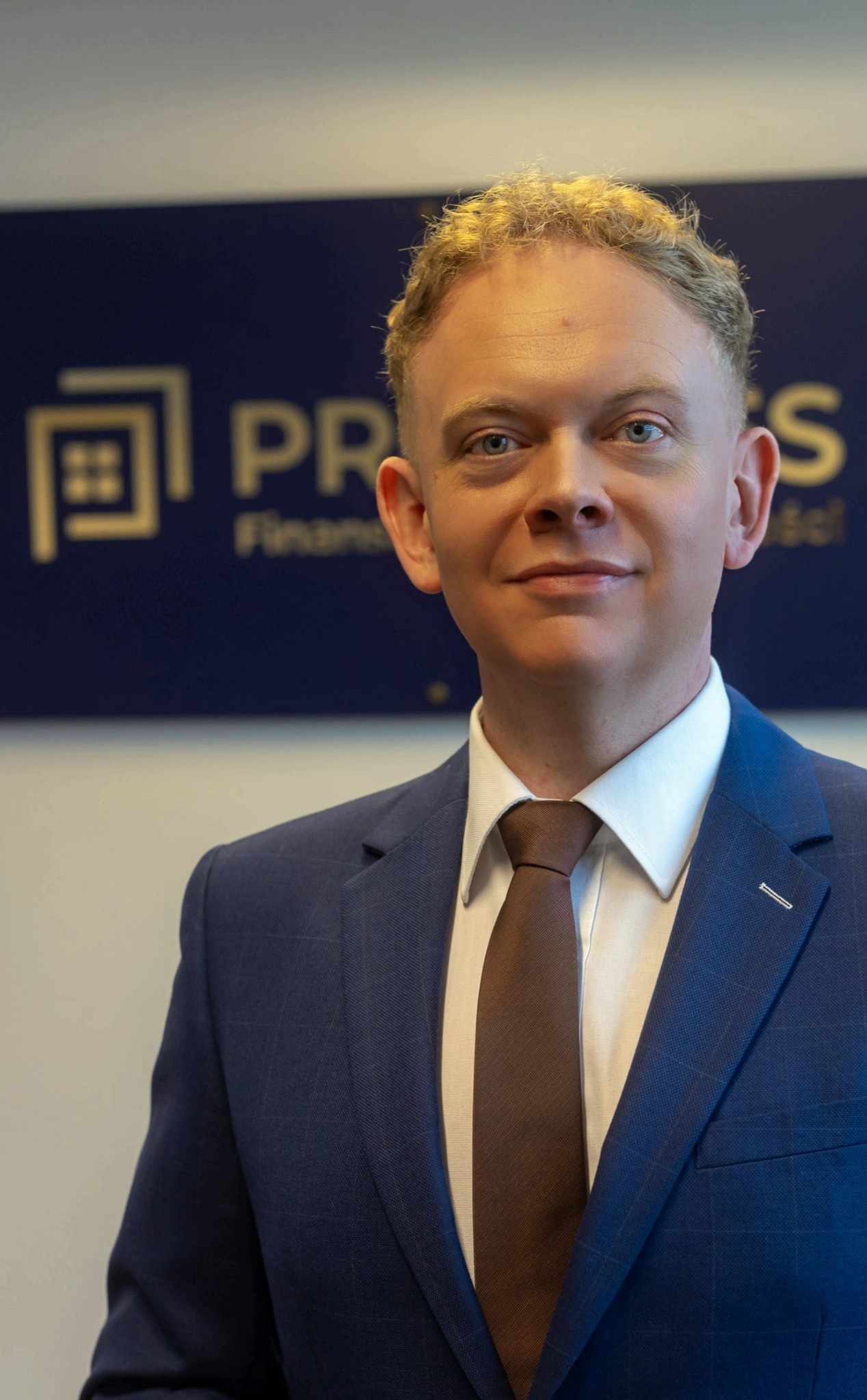 Portret uśmiechniętego mężczyzny w niebieskim garniturze i brązowym krawacie na tle logo firmy finansowej z motywem domu.