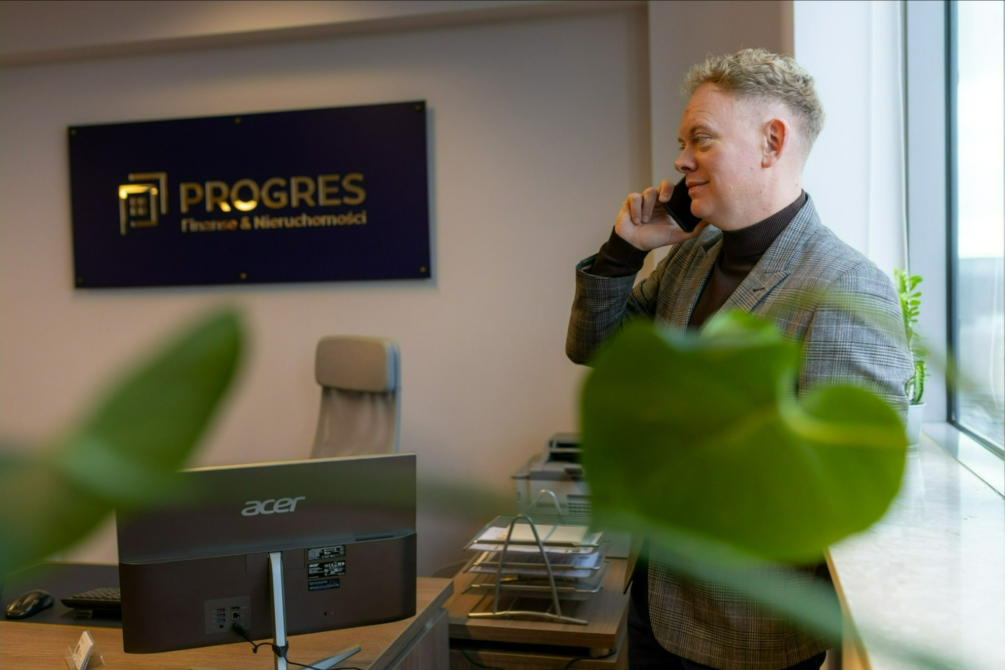 Mężczyzna w biurze rozmawia przez telefon, w tle logo firmy finansowej i nieruchomości, widok zza rośliny doniczkowej.