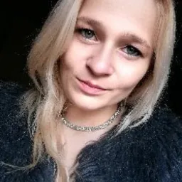 Portret kobiety z blond włosami i srebrnym naszyjnikiem, ubrana w futrzaną kamizelkę, spoglądająca w obiektyw.