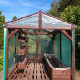 Drewniana konstrukcja szklarni ogrodowej z przezroczystym pokryciem, widoczne drewniane skrzynki na rośliny wewnątrz, podłoga z palet, w tle drzewa.