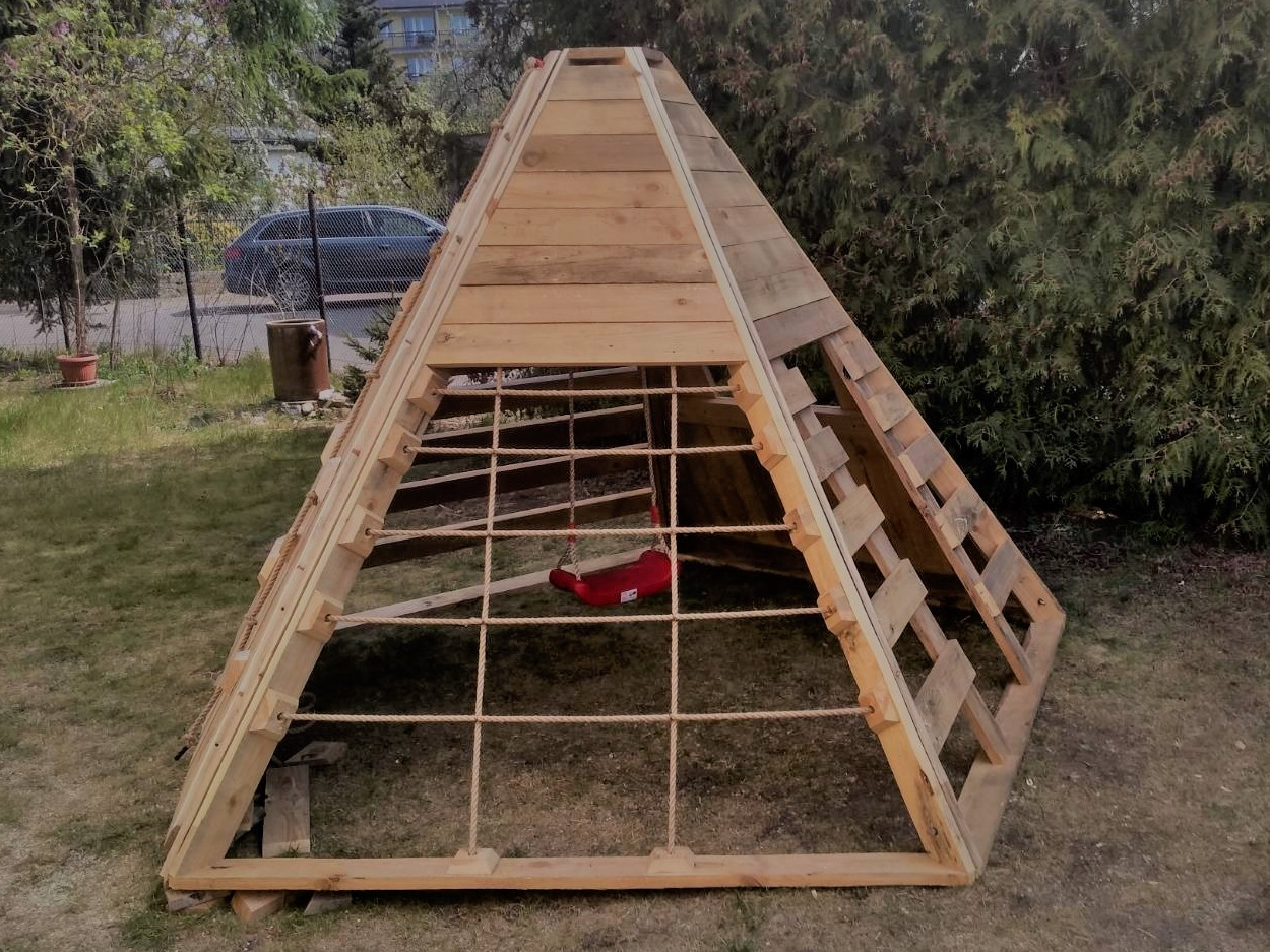 Domek ogrodowy dla dzieci w kształcie piramidy, wykonany z surowego drewna, z siatką sznurkową i czerwoną huśtawką wewnątrz, na tle zielonego trawnika i drzew.