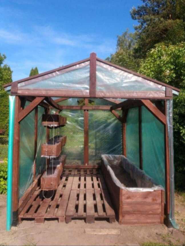 Drewniana konstrukcja szklarni ogrodowej z przezroczystym pokryciem, widoczne drewniane skrzynki na rośliny wewnątrz, podłoga z palet, w tle drzewa.