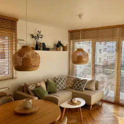 Jasne drewniane żaluzje w salonie z beżową sofą i drewnianym parkietem ułożonym w jodełkę, oświetlone ciepłym światłem słonecznym.