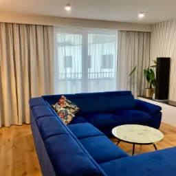 Eleganckie beżowe zasłony panelowe w nowoczesnym salonie z niebieską sofą narożną i stolikiem kawowym z marmurowym blatem.