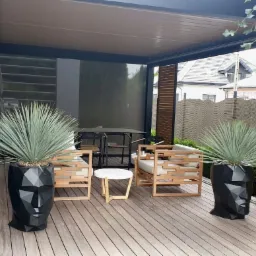 Nowoczesna pergola z drewnianą podłogą, meblami ogrodowymi i dekoracyjnymi osłonami przeciwsłonecznymi w kolorze beżowym, uzupełniona donicami w kształcie głowy.