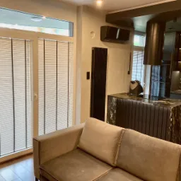 Nowoczesne wnętrze salonu z widokiem na okno balkonowe zabezpieczone białymi żaluzjami poziomymi z czarnymi sznurkami, obok beżowa sofa i wyspa kuchenna z czarnego marmuru.